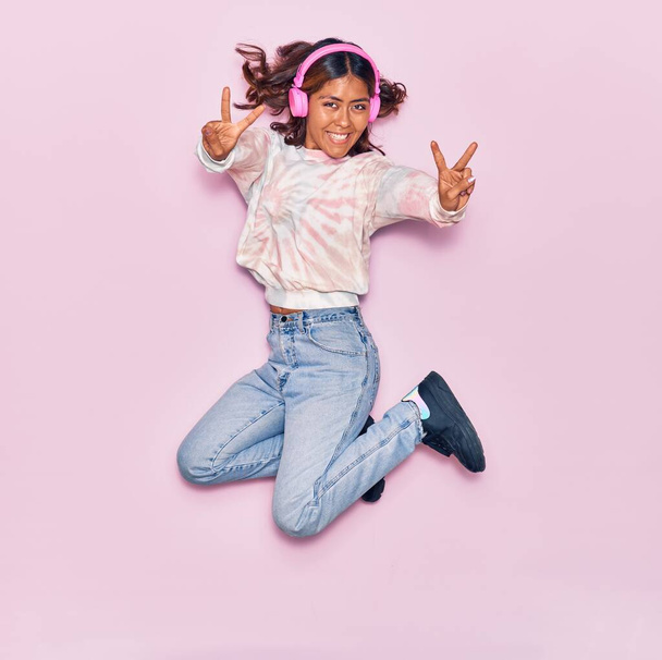 Joven hermosa mujer latina escuchando música usando auriculares sonriendo feliz. Saltar con sonrisa en la cara haciendo signo de victoria sobre fondo rosa aislado - Foto, imagen