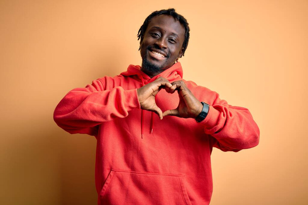 Jonge Afro-Amerikaanse sportieve man draagt sweatshirt met hoodie over gele achtergrond glimlachend in liefde doen hart symbool vorm met handen. Romantisch concept. - Foto, afbeelding