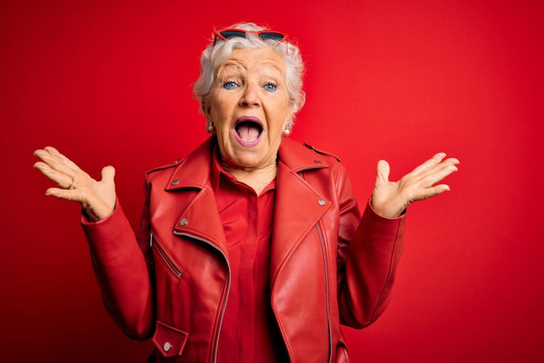 Senior beautiful grey-haired woman wearing casual red jacket and sunglasses celebrating crazy and amazed for success with arms raised and open eyes screaming excited. Winner concept - Foto, imagen