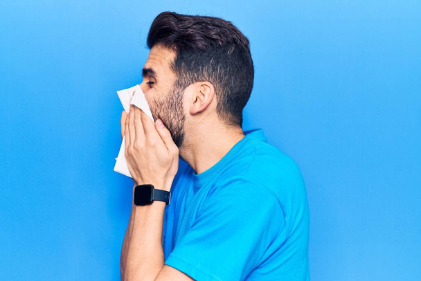 Jeune homme hispanique maladie utilisant mouchoir de papier sur le nez. Debout sur fond bleu isolé - Photo, image