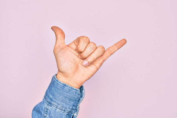 Hand of caucasian young man showing fingers over isolated pink background gesturing Hawaiian shaka greeting gesture, telephone and communication symbol - Foto, afbeelding