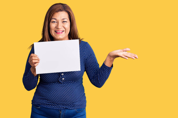 Donna latina di mezza età con striscione di carta con spazio vuoto che celebra la vittoria con sorriso felice e espressione del vincitore con le mani alzate  - Foto, immagini