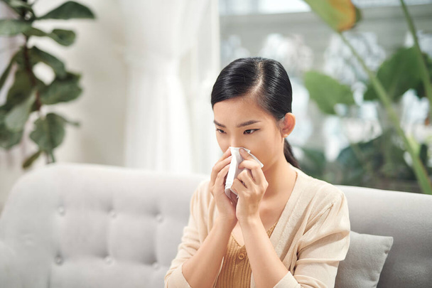 Mujer joven enferma sentada en el sofá sonándose la nariz en casa en la sala de estar. Foto de una mujer estornudando en papel. - Foto, Imagen
