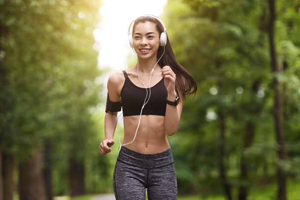 Ранковий біг. Усміхнена азіатська дівчина в навушниках бігає в зеленому парку
 - Фото, зображення