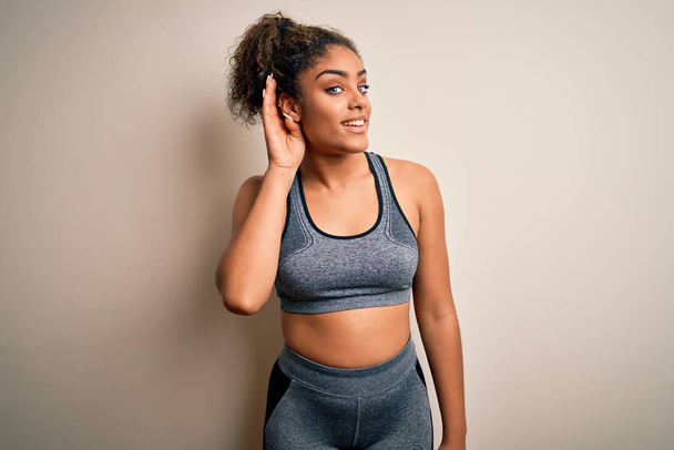 Young african american sportswoman doing sport wearing sportswear over white background smiling with hand over ear listening an hearing to rumor or gossip. Deafness concept. - Photo, Image
