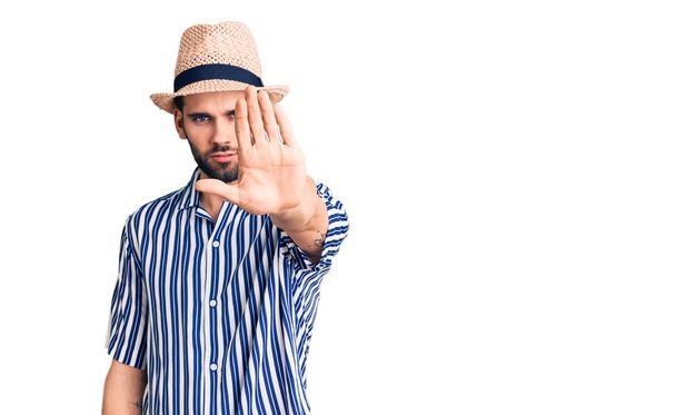 Young handsome man with beard wearing summer hat and striped shirt doing stop sing with palm of the hand. warning expression with negative and serious gesture on the face.  - Fotó, kép