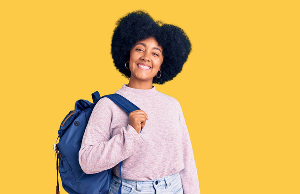 Молодая африканская американка с рюкзаком в руках выглядит позитивно и счастливо стоя и улыбаясь с уверенной улыбкой, показывая зубы  - Фото, изображение