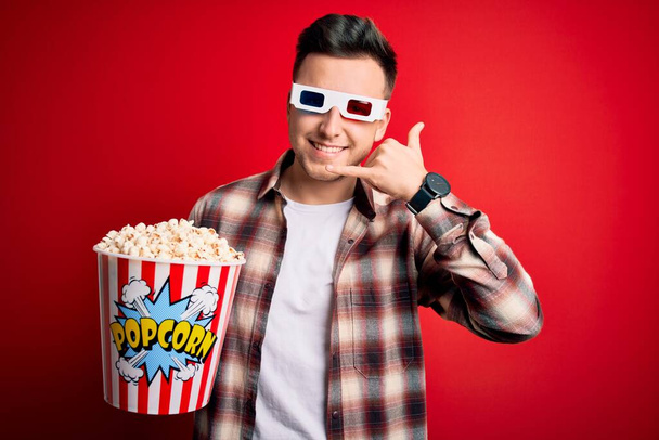 Молодий красивий кавказький чоловік носить 3d кіноокуляри і їсть попкорн, посміхаючись, роблячи телефонний жест рукою і пальцями, як розмовляти по телефону. комунікаційні концепції
. - Фото, зображення