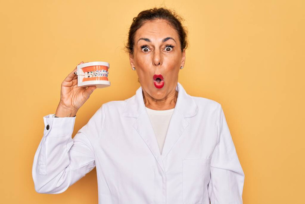 Старшая стоматолог среднего возраста женщина держит ортодонтию зубной протез с брекетами выравнивателя испуганный от шока с удивленным лицом, напуганный и взволнованный выражением страха - Фото, изображение