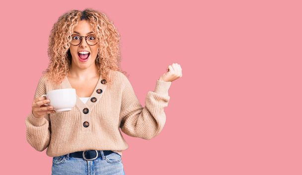 Young blonde woman with curly hair wearing glasses and drinking a cup of coffee screaming proud, celebrating victory and success very excited with raised arms  - Zdjęcie, obraz