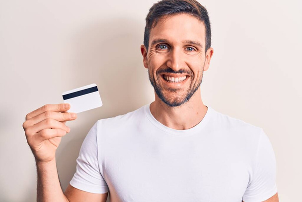 Молодой красивый клиент мужчина держит кредитную карту на изолированном белом фоне выглядит позитивно и счастливо стоя и улыбаясь с уверенной улыбкой показывая зубы - Фото, изображение