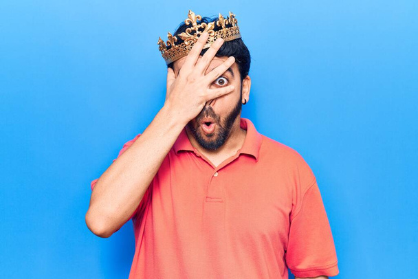 Young hispanic man wearing king crown peeking in shock covering face and eyes with hand, looking through fingers afraid  - Photo, Image