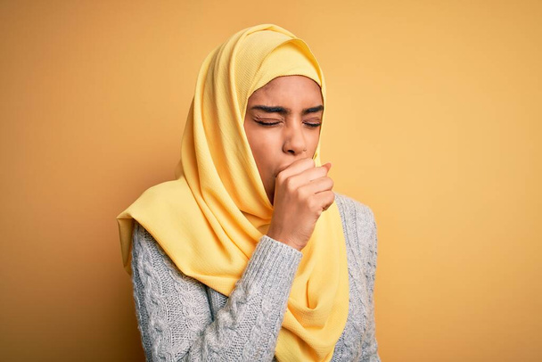 Mladá krásná africká americká dívka nosí muslimský hidžáb přes izolované žluté pozadí pocit nevolnosti a kašel jako symptom pro nachlazení nebo zánět průdušek. Koncept zdravotní péče. - Fotografie, Obrázek