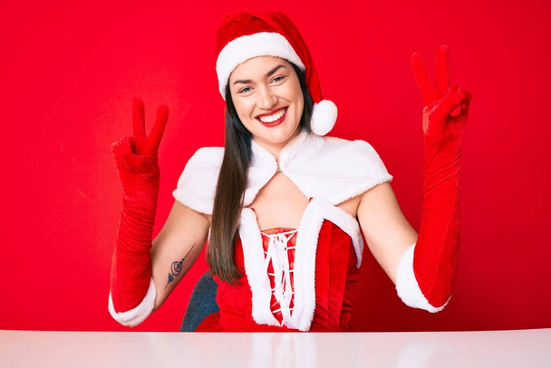 サンタの衣装を着た若い白人女性が勝利サインをしている指を見せてカメラを見て微笑んだ。2番.  - 写真・画像