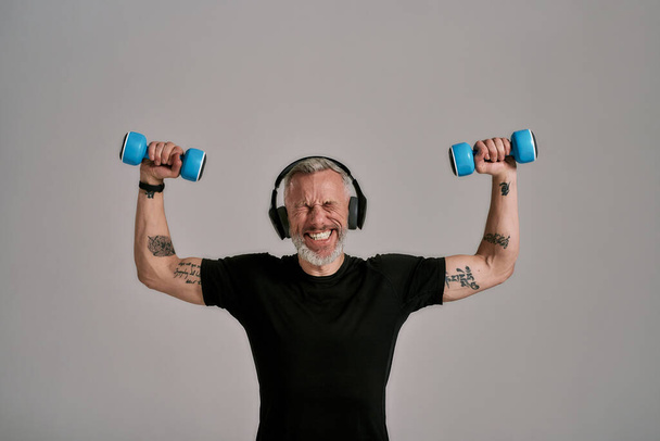 You can do it. Middle aged muscular man in black t shirt and headphones closing eyes, holding blue dumbbells, posing in studio over grey background - Foto, afbeelding