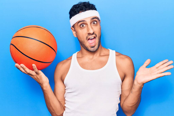 Young hispanic man holding basketball ball celebrating achievement with happy smile and winner expression with raised hand  - Zdjęcie, obraz