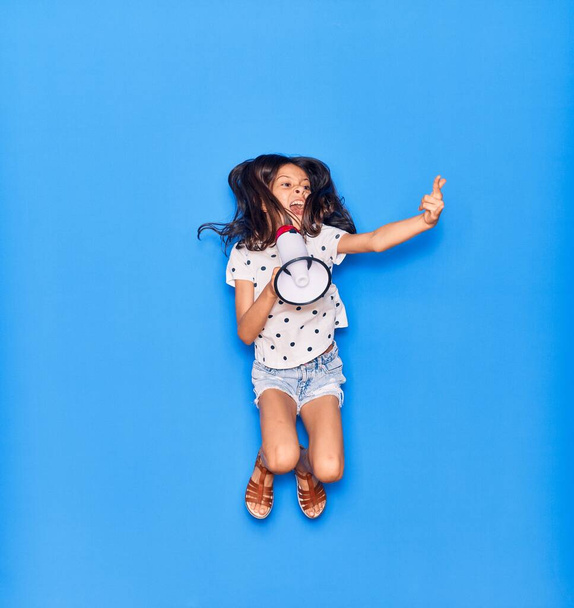 Rozkošná hispánská holčička křičící megafonem. Skákání dělá rohy znamení s prsty přes izolované modré pozadí - Fotografie, Obrázek