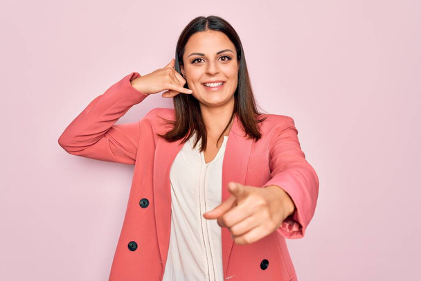 Young beautiful brunette businesswoman wearing elegant jacket over isolated pink background smiling doing talking on the telephone gesture and pointing to you. Call me. - Zdjęcie, obraz
