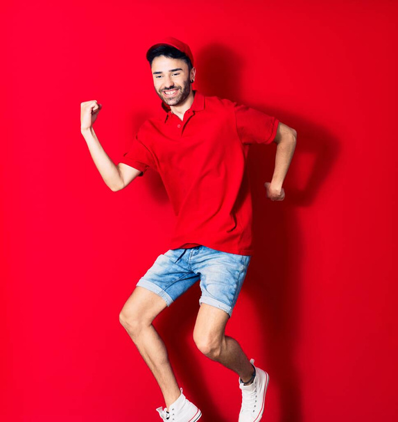 Young handsome deliveryman wearing cap smiling happy. Jumping with smile on face over isolated red background - Fotoğraf, Görsel