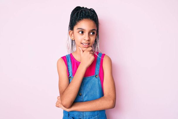 Νεαρό Αφροαμερικανό κορίτσι με κοτσίδες που φοράει καθημερινά ρούχα πάνω από ροζ φόντο με το χέρι στο πηγούνι να σκέφτεται την ερώτηση, την σκεπτική έκφραση. Χαμογελώντας με ευγενικό πρόσωπο. έννοια της αμφιβολίας.  - Φωτογραφία, εικόνα