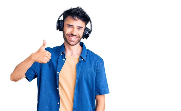 Joven hombre hispano escuchando música usando auriculares sonriendo feliz y positivo, pulgar hacia arriba haciendo excelente y signo de aprobación  - Foto, imagen