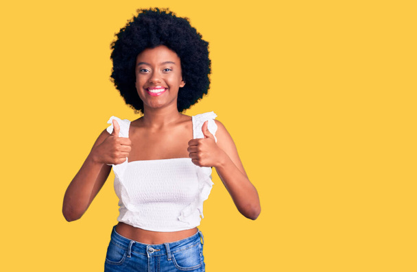 カジュアルな服を着ている若いアフリカ系アメリカ人女性は、手で積極的なジェスチャーを行い、笑顔と幸せを親指アップ。陽気な表情と勝者のジェスチャー.  - 写真・画像