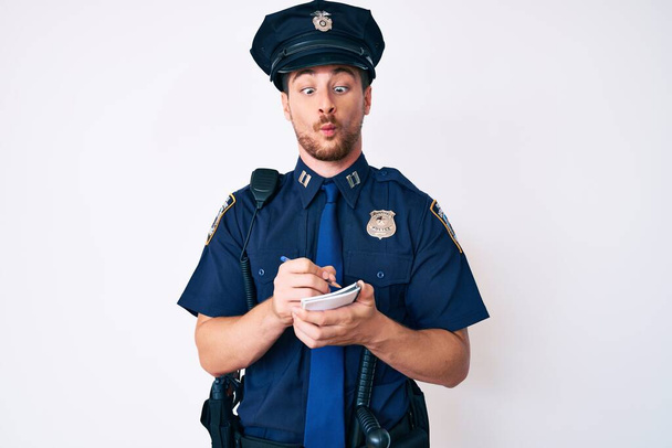 Jonge blanke man draagt politie-uniform schrijven prima maken vis gezicht met mond en squinting ogen, gek en komisch.  - Foto, afbeelding