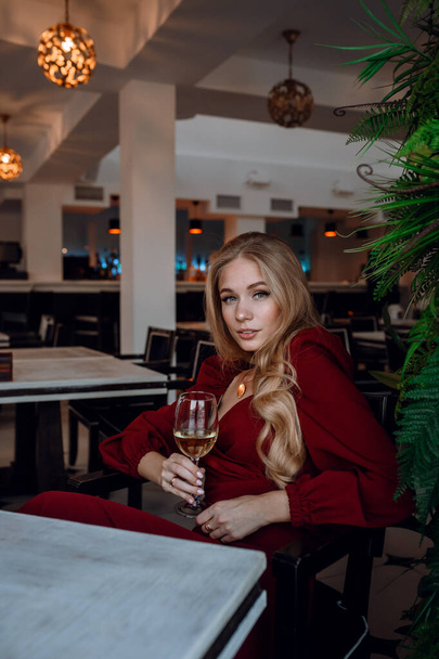 woman with glass of white wine in cafe or restaurant. sexy young blonde girl. concept of celebration, enjoying alcohol, style and fashionable lifestyle. Portrait girl in red dress with wine glass - Photo, image
