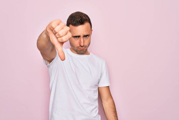 青い目のハンサムな男は、ピンクの背景に立ってカジュアルな白いTシャツを着て、不満と怒りを示し拒絶反応と親指ダウンジェスチャーで陰性に見えます。悪い表現. - 写真・画像