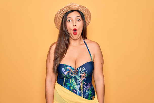 Jonge Spaanse vrouw met zomerhoed en badpak over gele achtergrond bang en geschokt met verrassingsexpressie, angst en opgewonden gezicht. - Foto, afbeelding