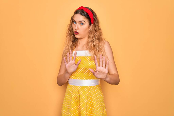 Beautiful blonde pin-up woman with blue eyes wearing diadem standing over yellow background Moving away hands palms showing refusal and denial with afraid and disgusting expression. Stop and forbidden. - Foto, Bild