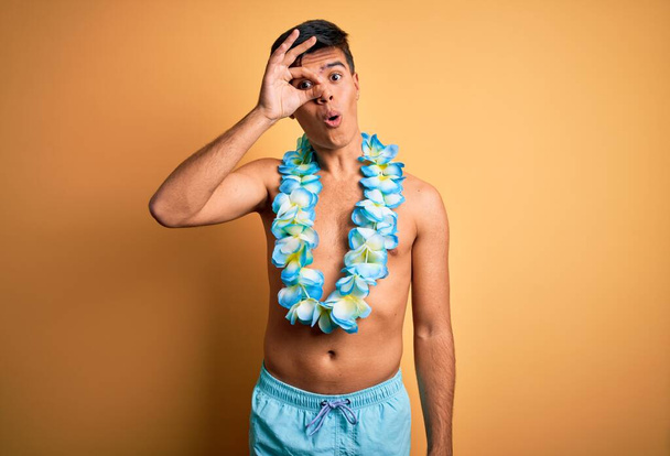 Joven hombre guapo turista de vacaciones con trajes de baño y flores de lei hawaiano haciendo el gesto bien sorprendido con la cara sorprendida, los ojos mirando a través de los dedos. Expresión incrédula
. - Foto, Imagen