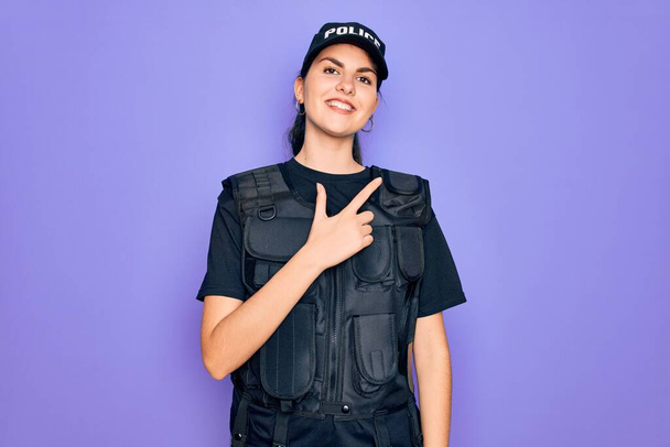 Νεαρή αστυνομικός φορώντας αλεξίσφαιρο γιλέκο ασφαλείας στολή πάνω από μωβ φόντο χαρούμενα με ένα χαμόγελο στο πρόσωπο δείχνει με το χέρι και το δάχτυλο μέχρι το πλάι με ευτυχισμένη και φυσική έκφραση - Φωτογραφία, εικόνα