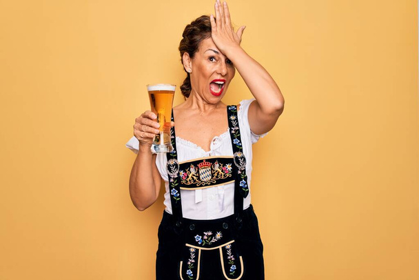 Μέση ηλικία μελαχρινή γυναίκα φορώντας γερμανικό παραδοσιακό φόρεμα Oktoberfest πίνοντας ποτήρι μπύρα τόνισε με το χέρι στο κεφάλι, σοκαρισμένος από ντροπή και έκπληξη πρόσωπο, θυμωμένος και απογοητευμένος. Φόβος και αναστάτωση για λάθος. - Φωτογραφία, εικόνα