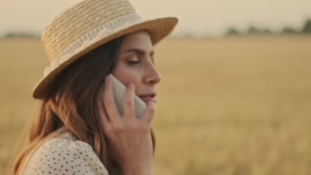 Close up view of Happy attractive woman wearing dress and stylish hat talking by smartphone while walking on wheat field - Filmati, video