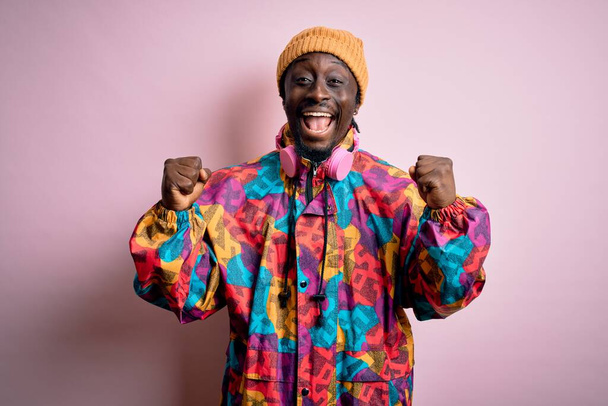 Νεαρός όμορφος Αφροαμερικάνος με πολύχρωμο παλτό και καπέλο πάνω από ροζ φόντο γιορτάζει έκπληκτος και έκπληκτος για την επιτυχία με τα χέρια ψηλά και ανοιχτά μάτια. Έννοια νικητή. - Φωτογραφία, εικόνα