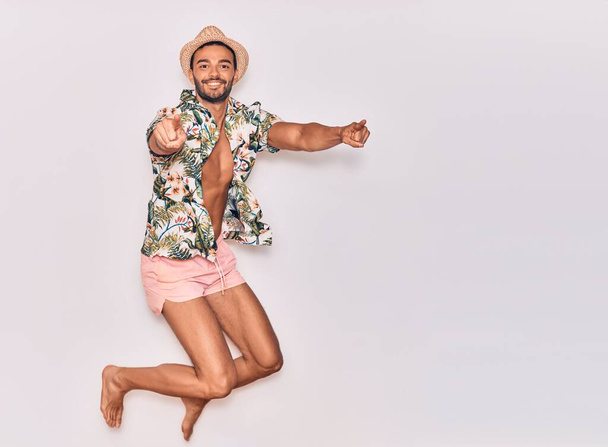 Joven hombre hispano guapo de vacaciones vistiendo trajes de baño, camisa floral y sombrero sonriendo feliz. Saltando con sonrisa en la cara señalando con los dedos sobre el fondo blanco aislado - Foto, Imagen