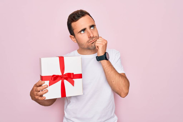 Молодой красивый мужчина с голубыми глазами держит подарок на день рождения на изолированном розовом фоне серьезное лицо, думая о вопросе, очень запутанная идея - Фото, изображение