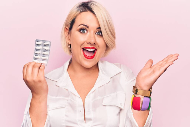 Young beautiful blonde plus size woman holding pills over isolated pink background celebrating achievement with happy smile and winner expression with raised hand - Foto, Bild