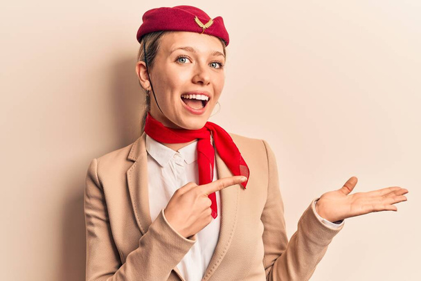 Młoda piękna blondynka w mundurze stewardessy zdumiona i uśmiechnięta do aparatu prezentując się z ręką i wskazując palcem.  - Zdjęcie, obraz