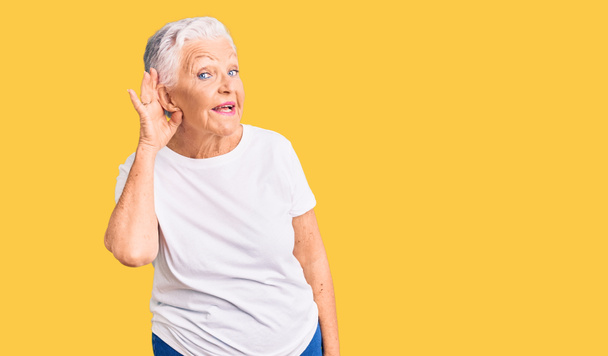 Belle femme âgée aux yeux bleus et aux cheveux gris portant un t-shirt blanc décontracté souriant main sur oreille en écoutant une rumeur ou des commérages. concept de surdité.  - Photo, image