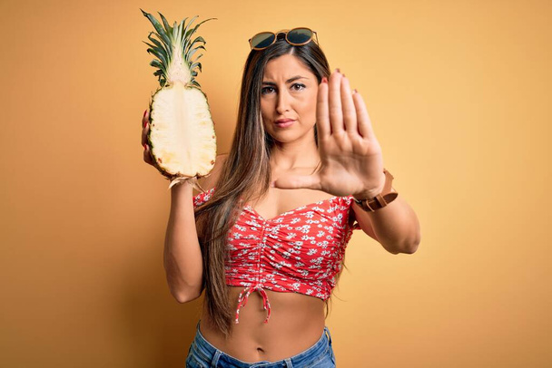 Młoda piękna brunetka trzyma środkowy owoc ananasa na odizolowanym żółtym tle z otwartą ręką robi znak stop z poważnym i pewnym siebie wyrazem, gest obrony - Zdjęcie, obraz