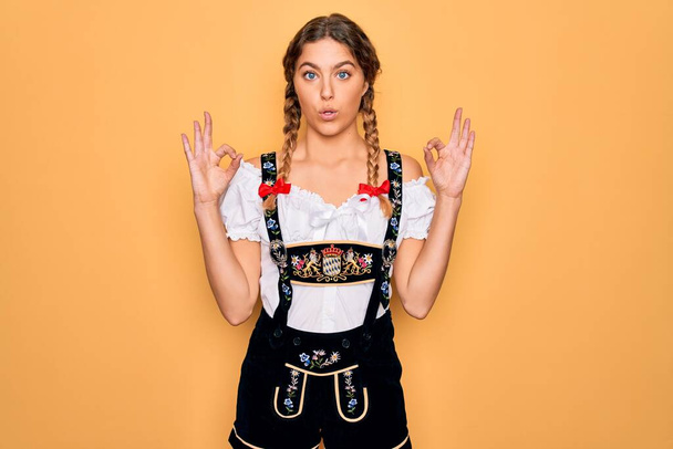 Young beautiful blonde german woman with blue eyes wearing traditional octoberfest dress looking surprised and shocked doing ok approval symbol with fingers. Crazy expression - Photo, Image