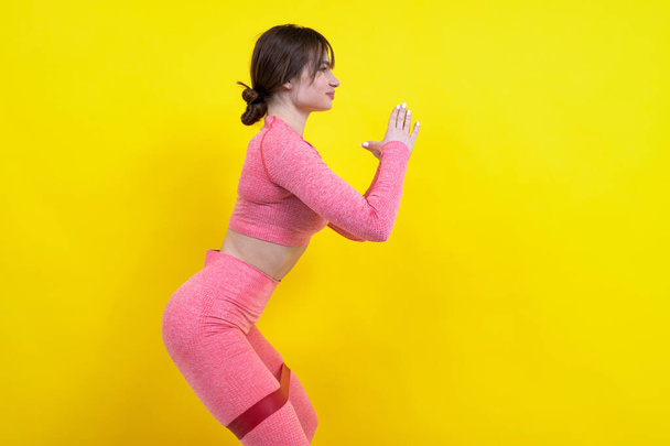 A young girl in a pink tracksuit trains with a fitness elastic band on a yellow background - Фото, изображение