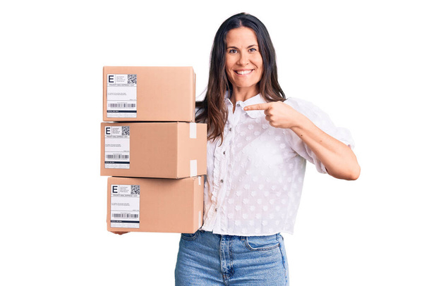 Young beautiful brunette woman holding delivery cardboard package pointing finger to one self smiling happy and proud  - Photo, Image