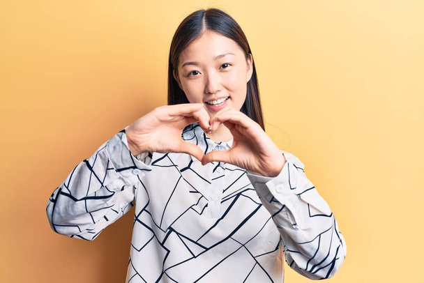 Giovane bella donna cinese indossa camicia elegante sorridente in amore facendo forma di simbolo del cuore con le mani. concetto romantico.  - Foto, immagini