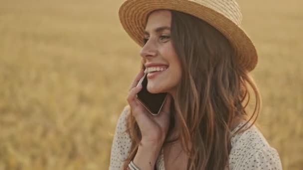 Happy attractive woman wearing dress and stylish hat talking by smartphone and laughing while walking on wheat field - Filmati, video