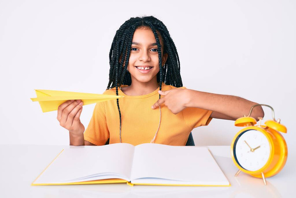 Νεαρό Αφροαμερικανό κορίτσι με κοτσίδες να κρατάει χάρτινο αεροπλάνο ενώ μελετούσε δείχνοντας τον εαυτό του χαμογελώντας χαρούμενο και περήφανο.  - Φωτογραφία, εικόνα