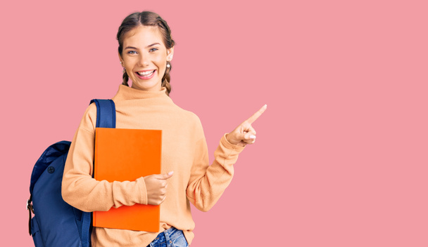 Beautiful caucasian woman with blonde hair wearing student backpack and holding book smiling happy pointing with hand and finger to the side  - Photo, image