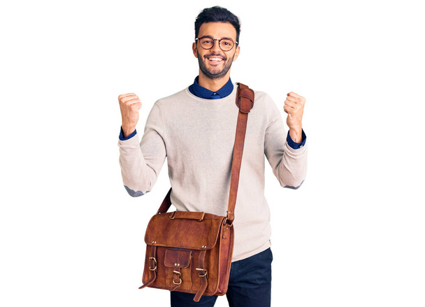 Young handsome hispanic man wearing leather bag screaming proud, celebrating victory and success very excited with raised arms  - Foto, imagen